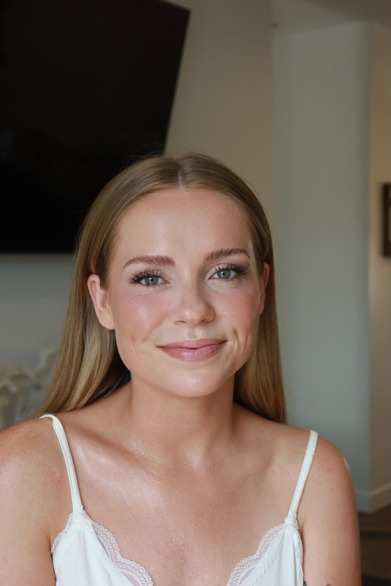 A blonde woman in a white camisole with sleek hair and soft glam bridal makeup, looking at the camera