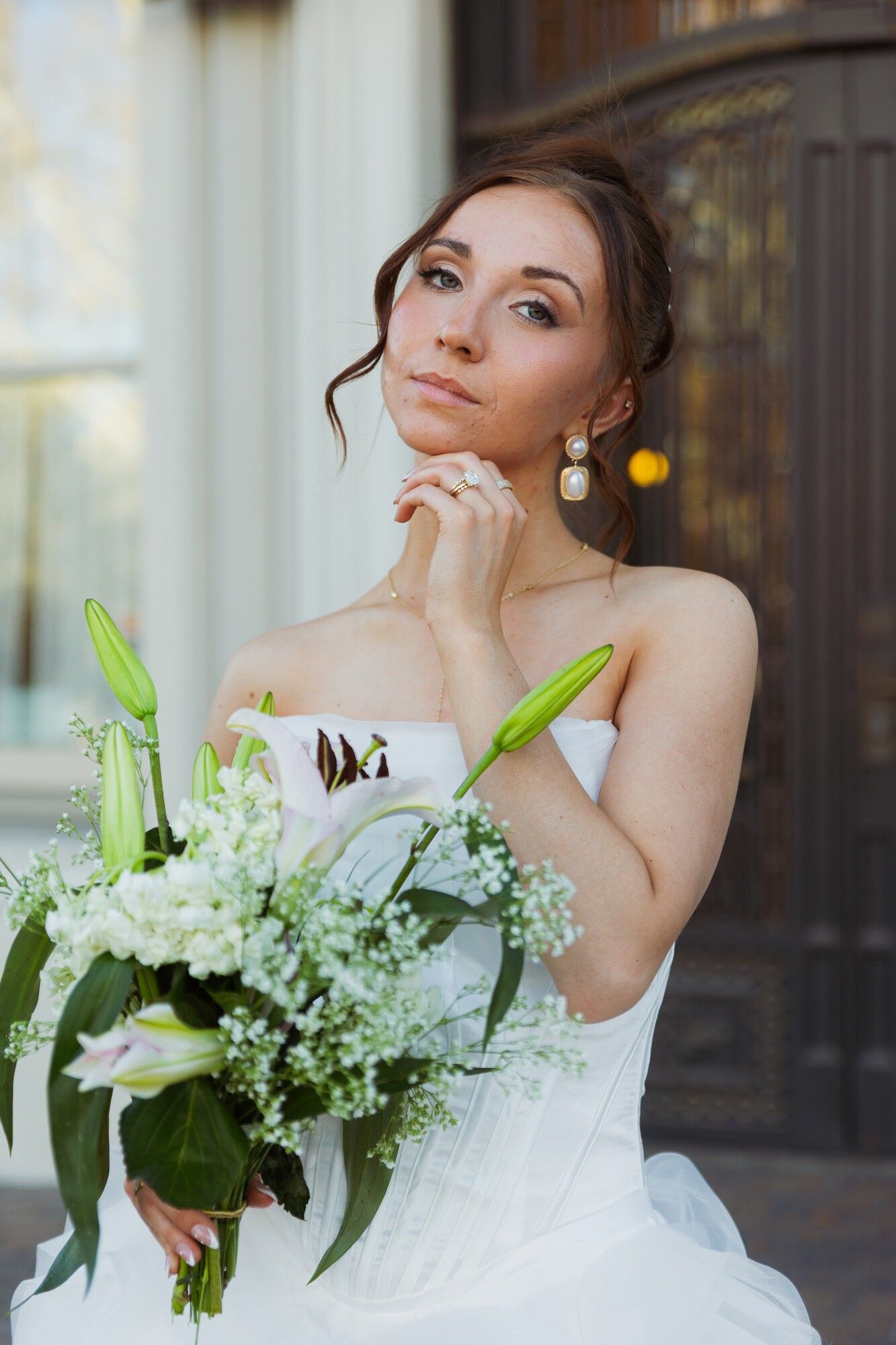 A bride holding a white bouquet with lilies, hand to her chin, pearl earrings and a soft updo