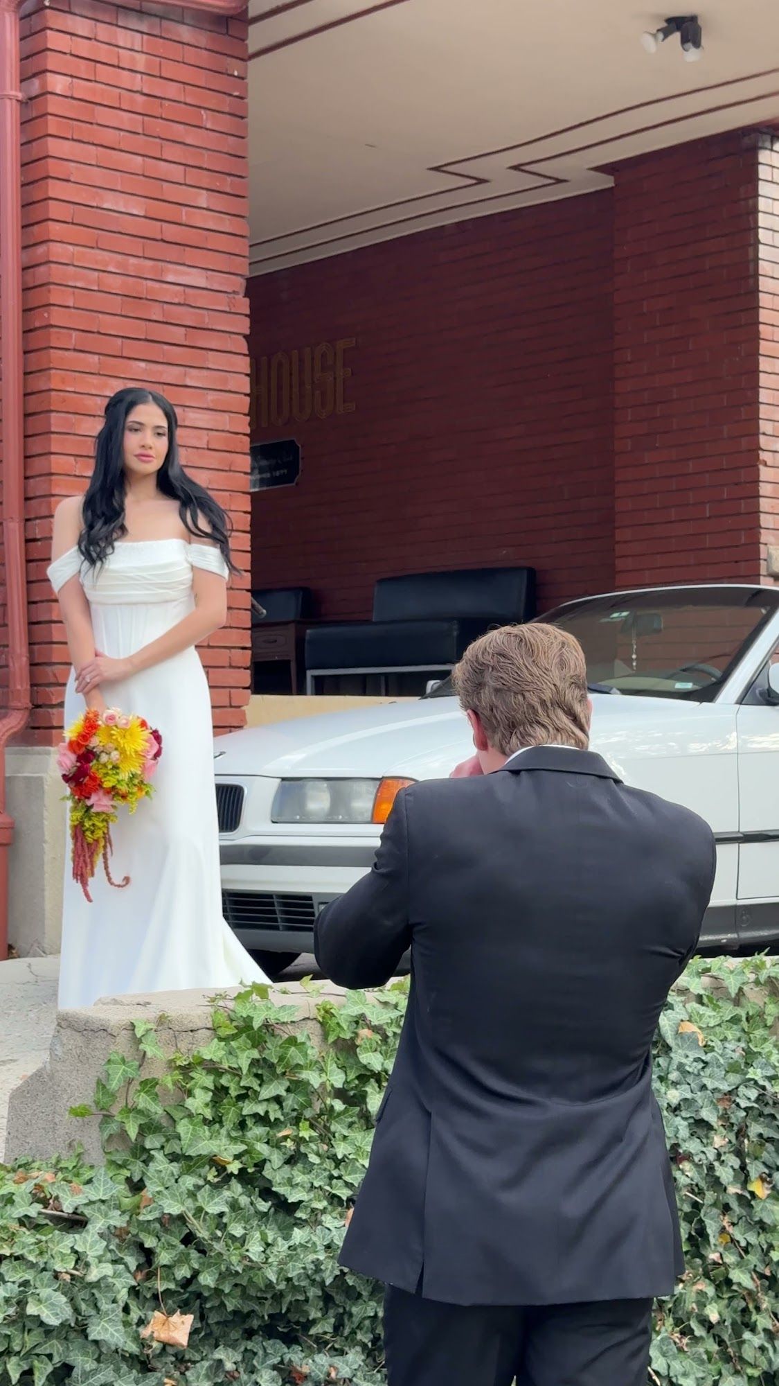 A bride in a simple white gown holding a red bouquet outside a brick building, her groom and a white classic car beside her