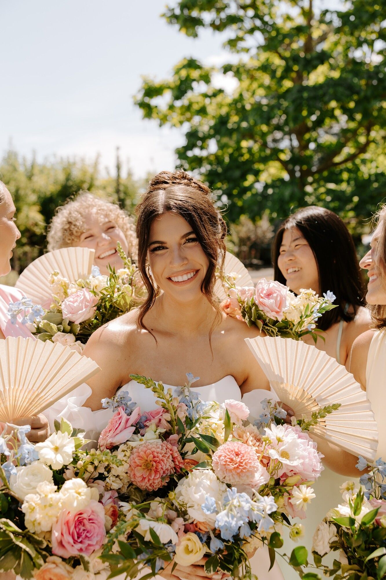 A bride surrounded by bridesmaids holding paper fans and pastel bouquets in the sun