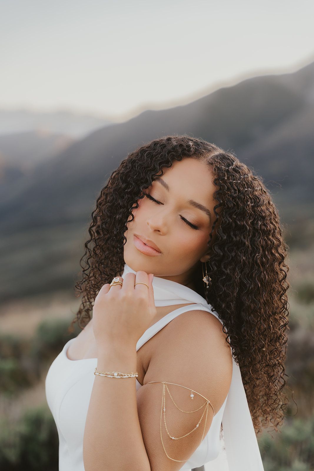 A bride with long natural curls and soft glam makeup, eyes closed, mountains behind her
