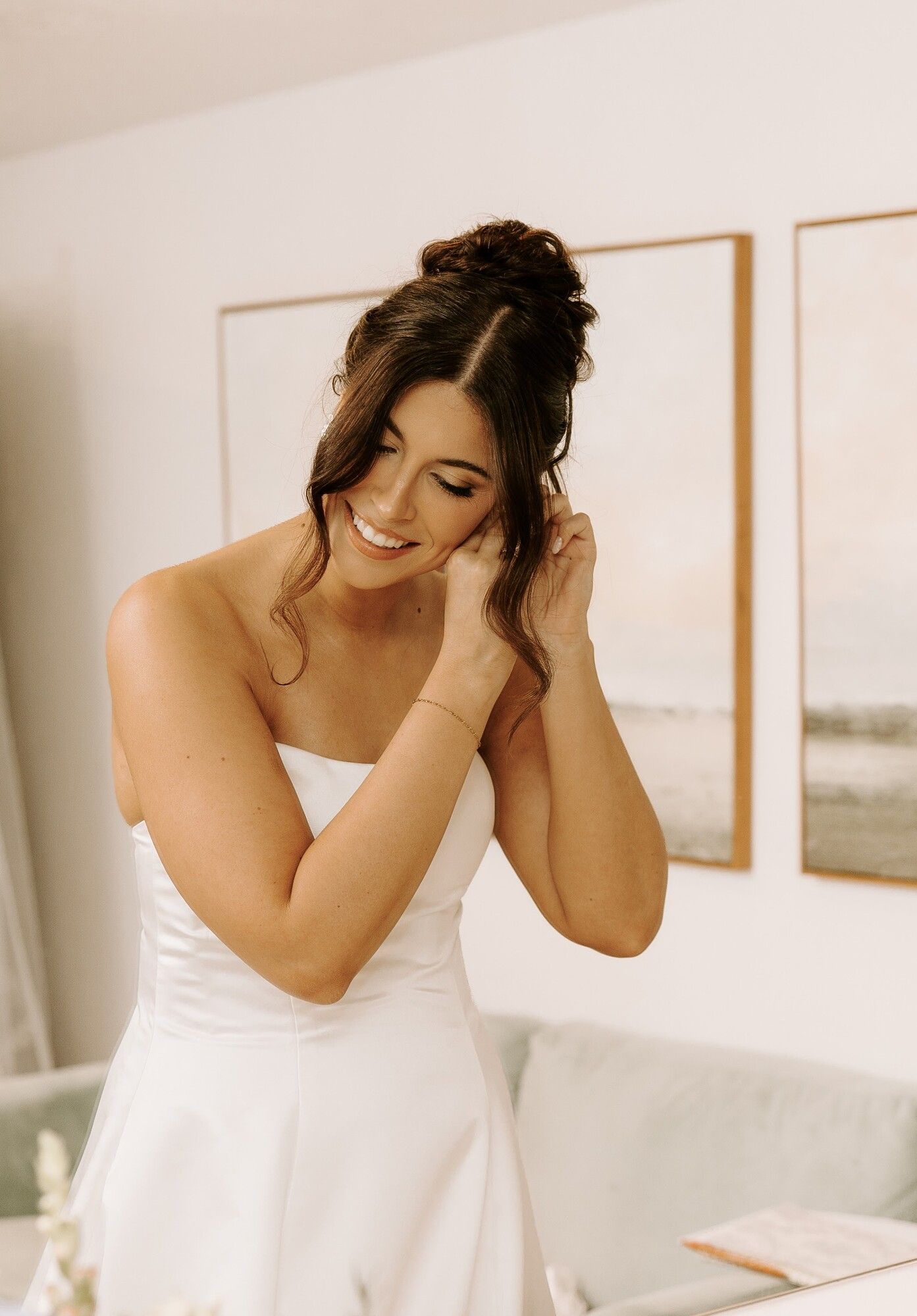 A bride in a white gown putting on an earring, laughing, in a bright getting-ready room