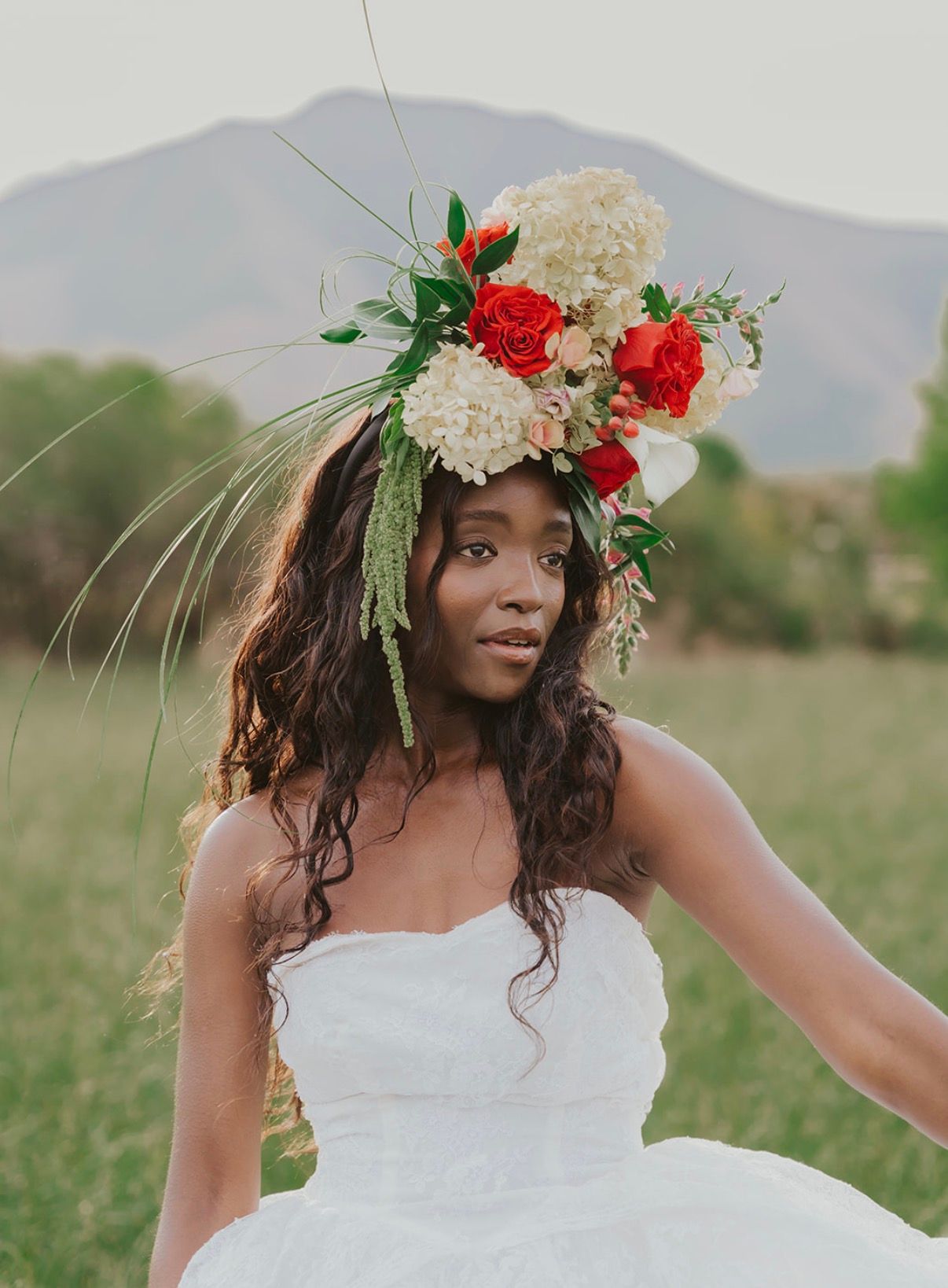 A bride in a full white gown and a large red and white flower crown in a mountain meadow