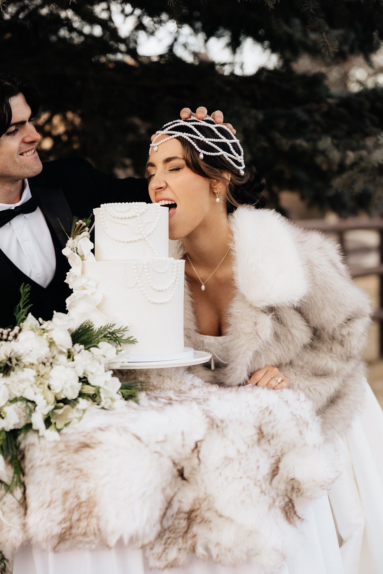 A bride in a fur wrap and pearl headpiece leaning in to bite her wedding cake outdoors