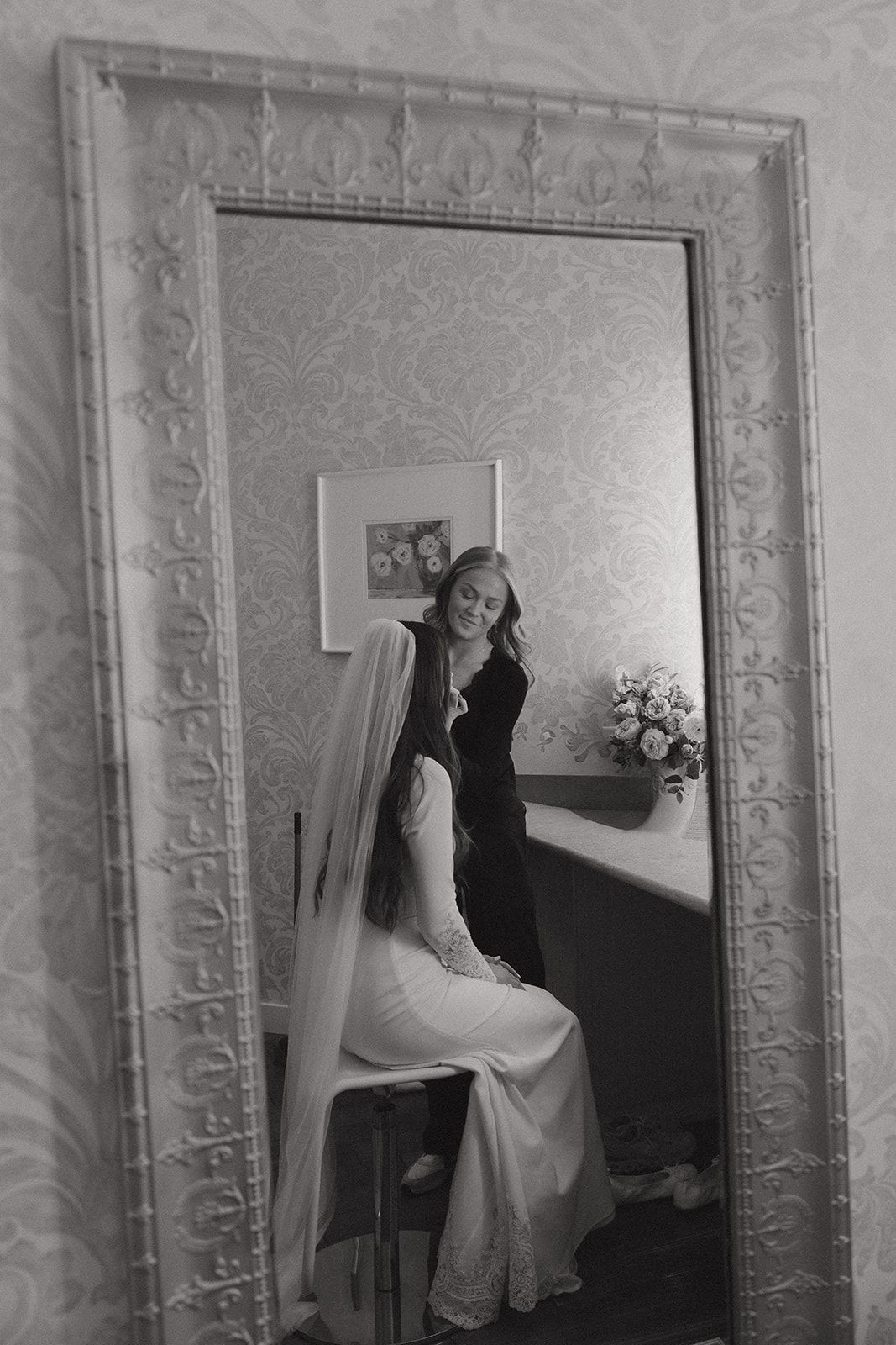A bride in her veil reflected in an ornate mirror, in black and white