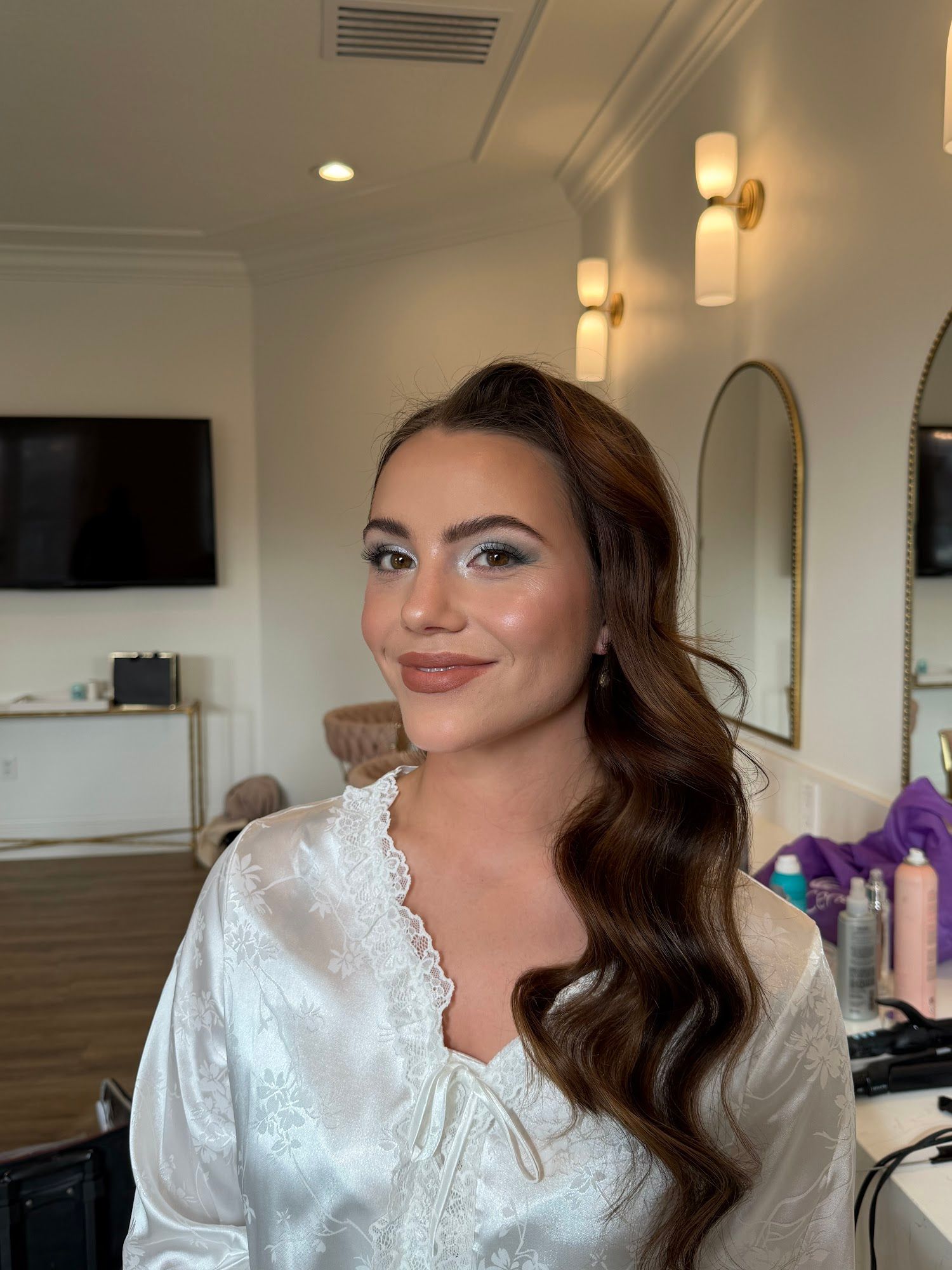 A dark-haired bride in a white satin robe with soft waves and finished makeup, in the getting-ready room