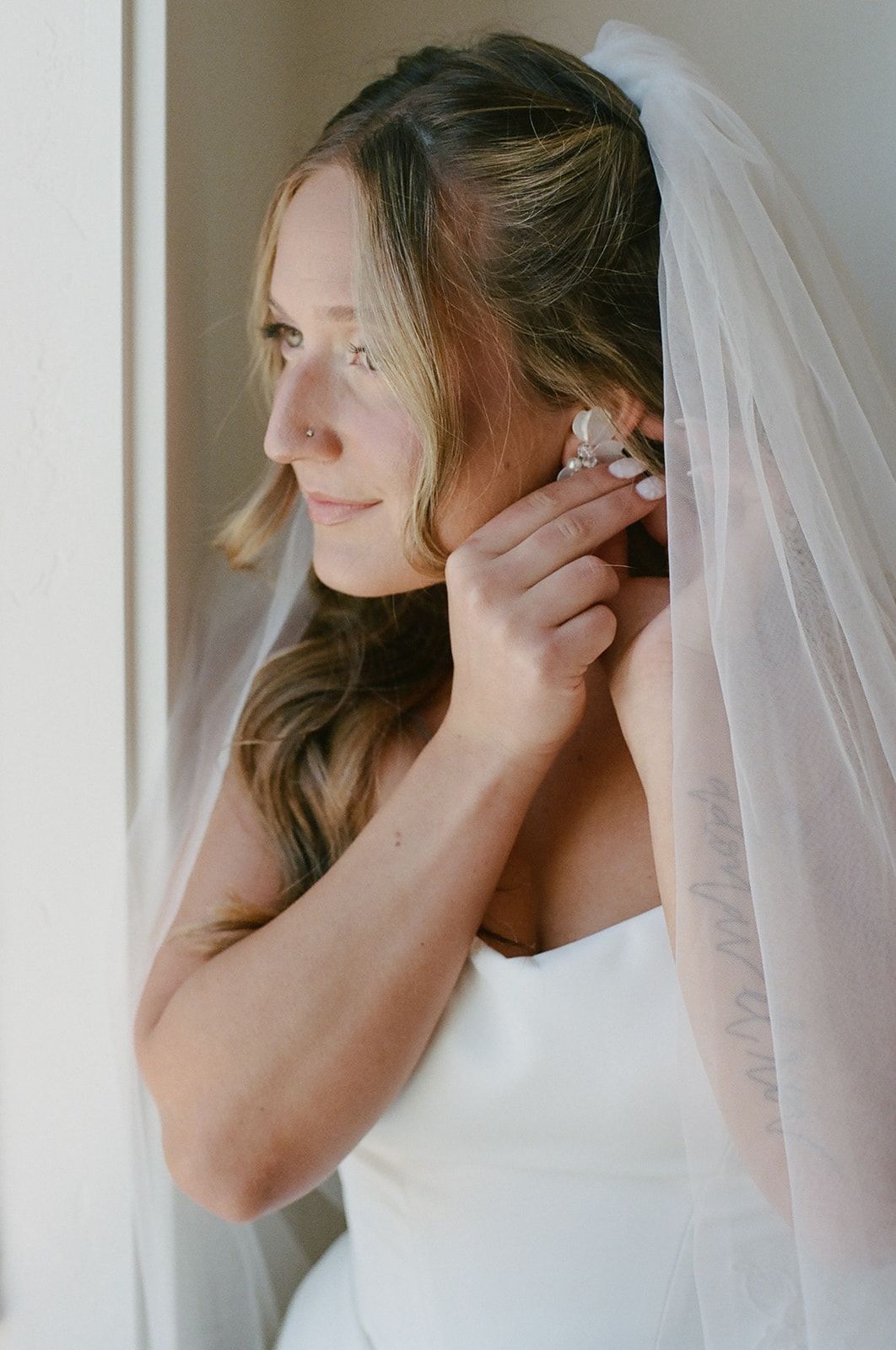 A bride in her veil fastening an earring by a bright window, soft glam makeup