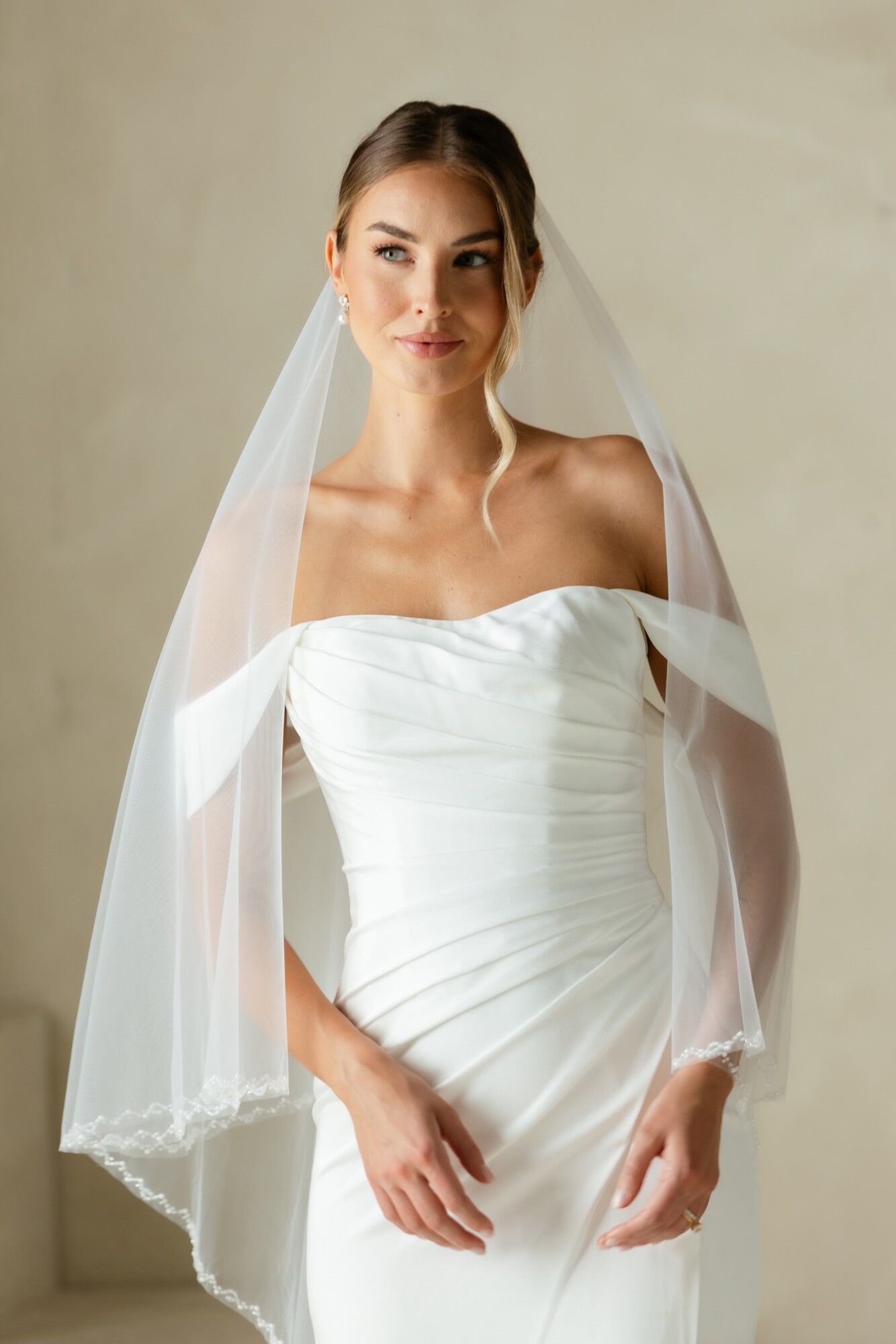 A bride standing in a draped white gown and long lace veil, hands clasped