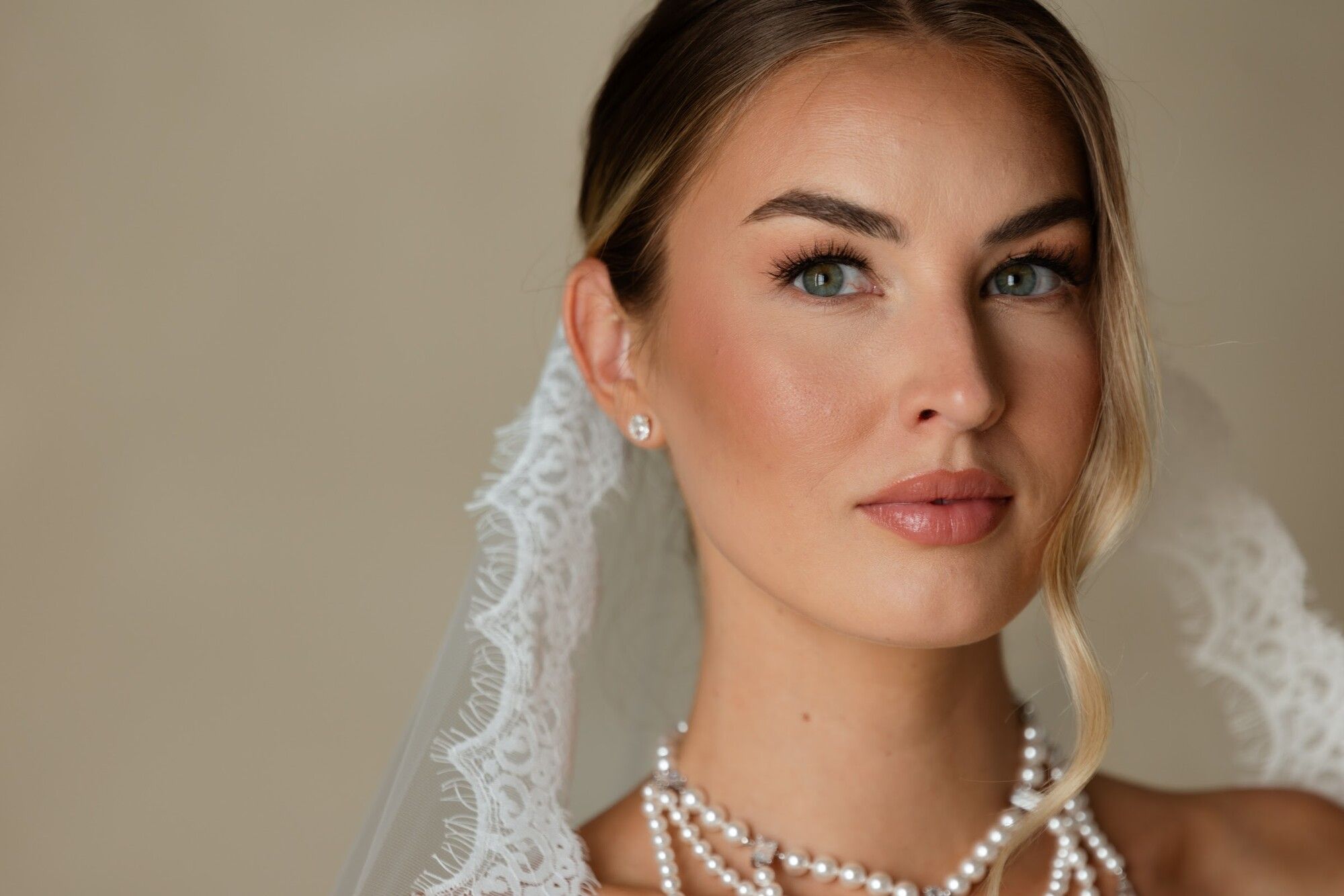 A bride in a lace-edged veil and layered pearls, soft glam makeup, in a warm room