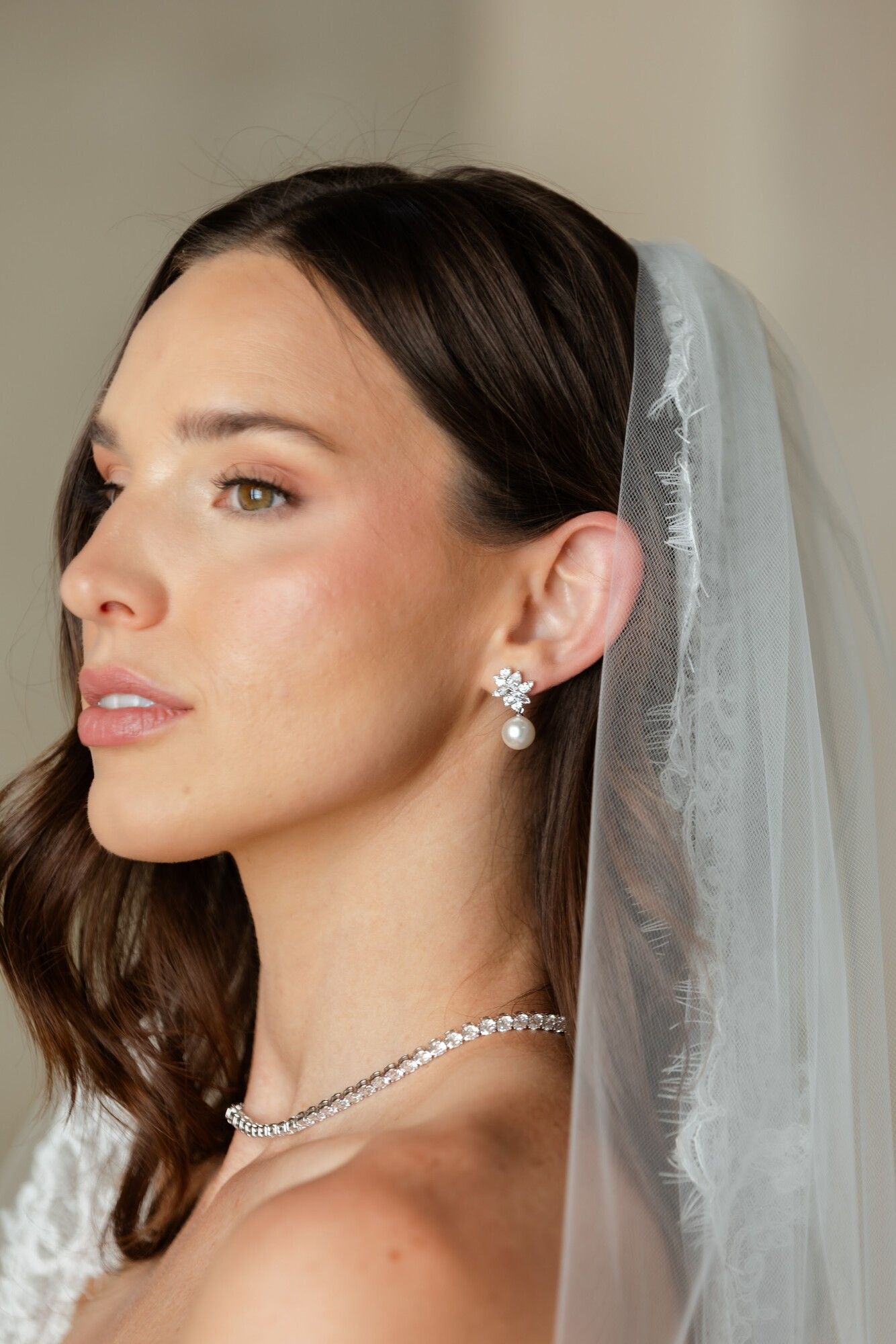 A bride in profile adjusting her lace veil, a diamond necklace and soft updo