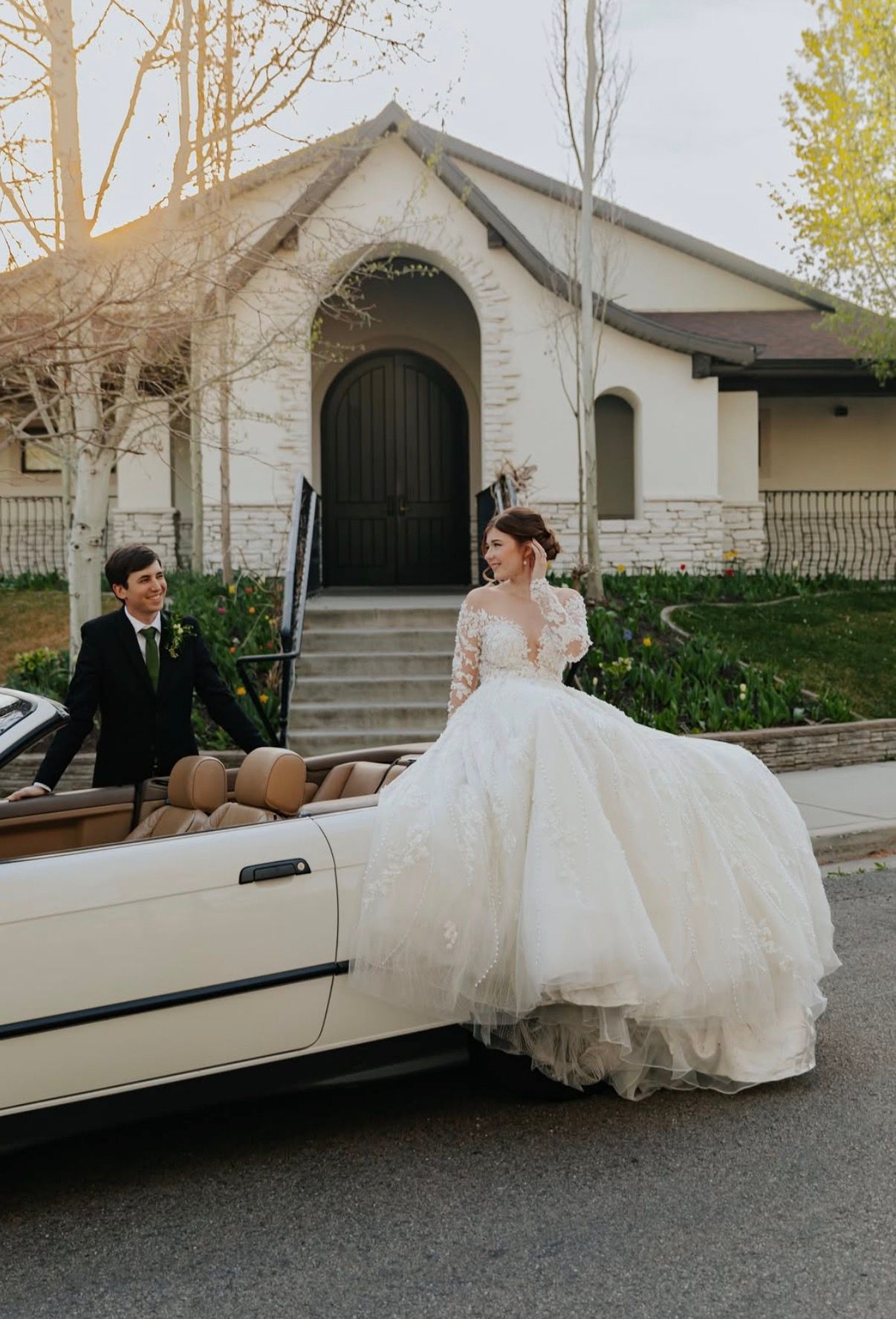 A bride in a full tulle gown sitting on the hood of a vintage car outside a white chapel, her groom at the wheel