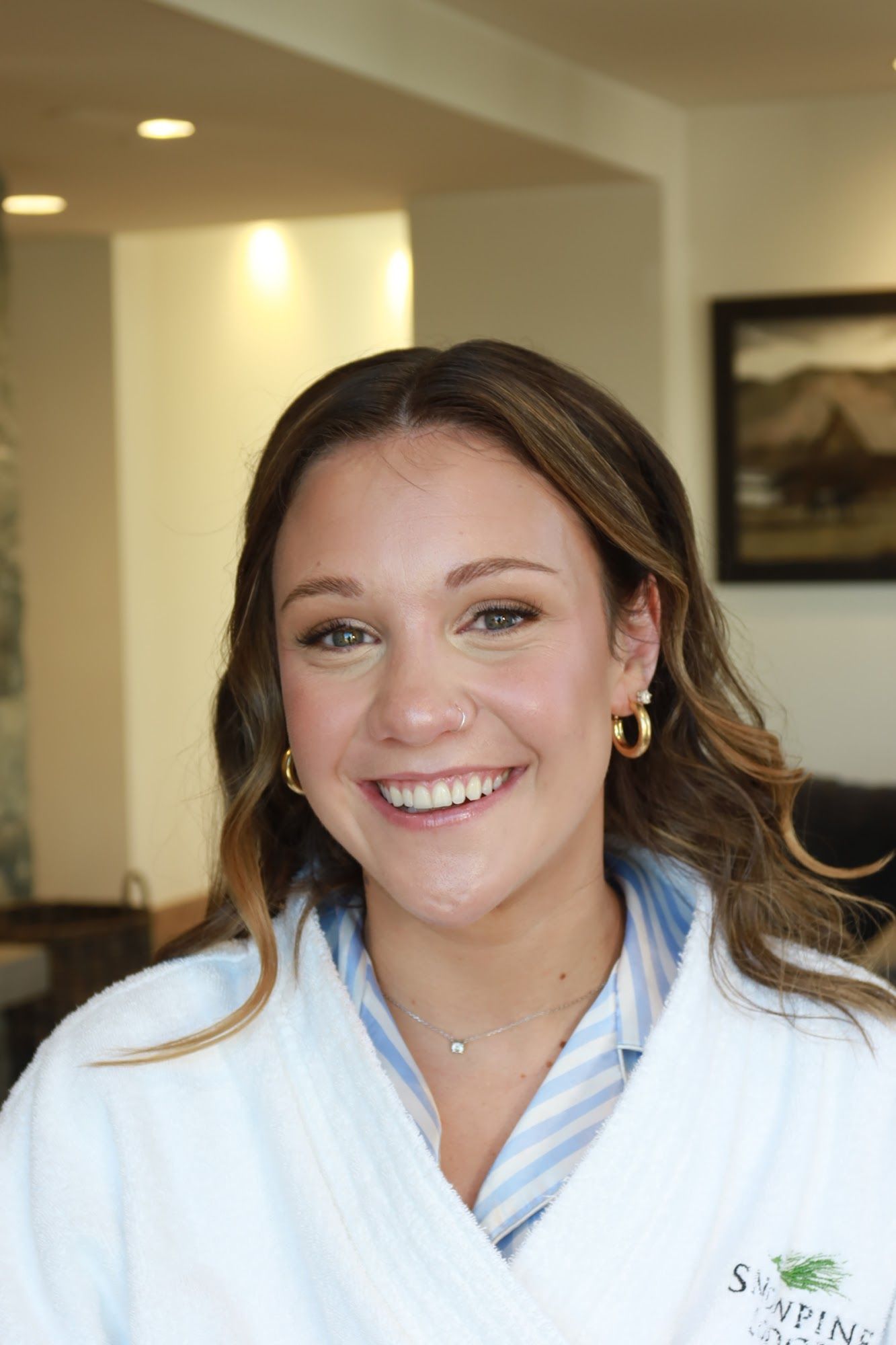 A bride in a white robe with soft waves and finished makeup, smiling in the getting-ready room at Snowpine Lodge