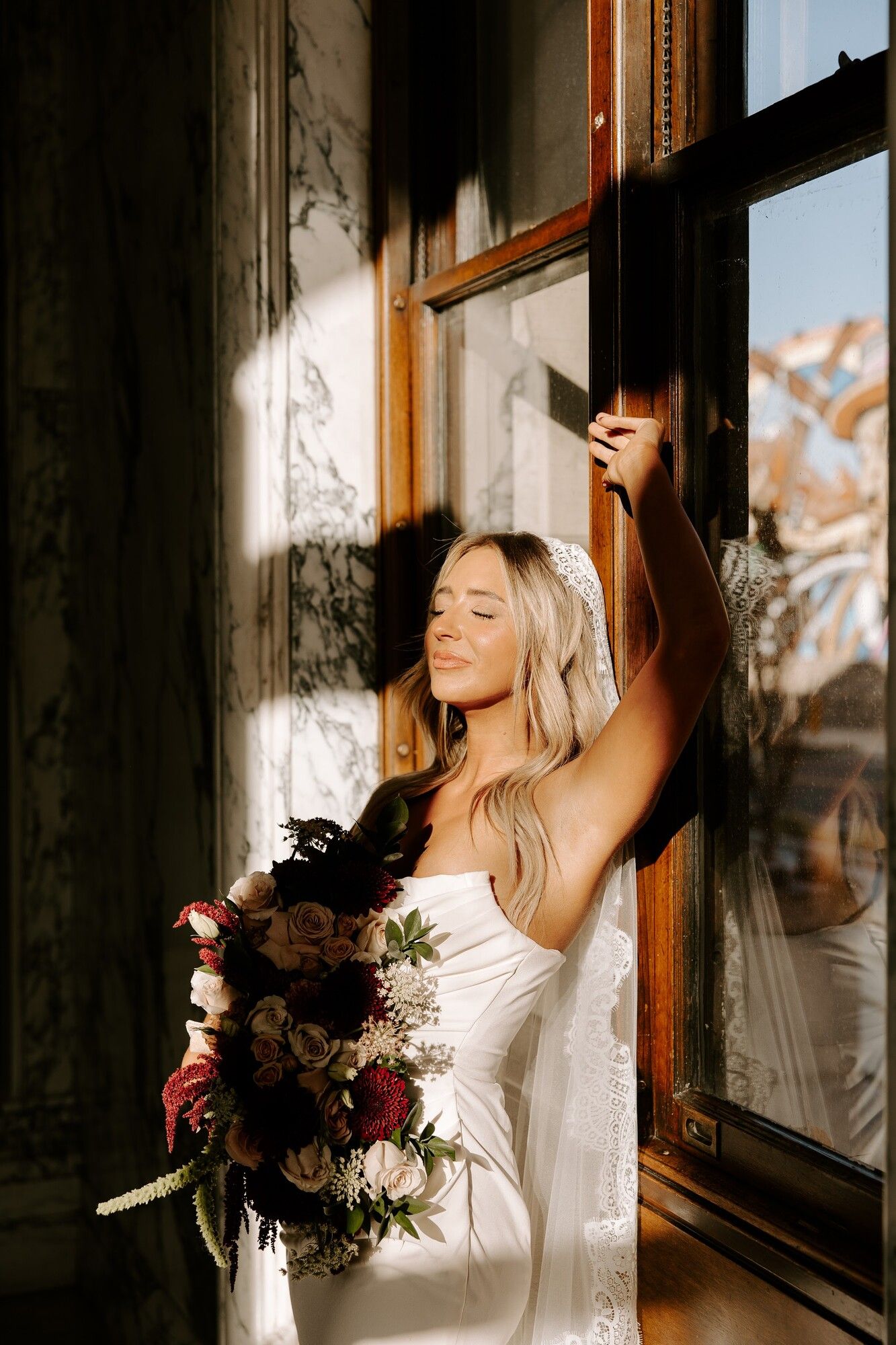 A bride in a sunlit window with a dark red bouquet, in a wood-panelled room