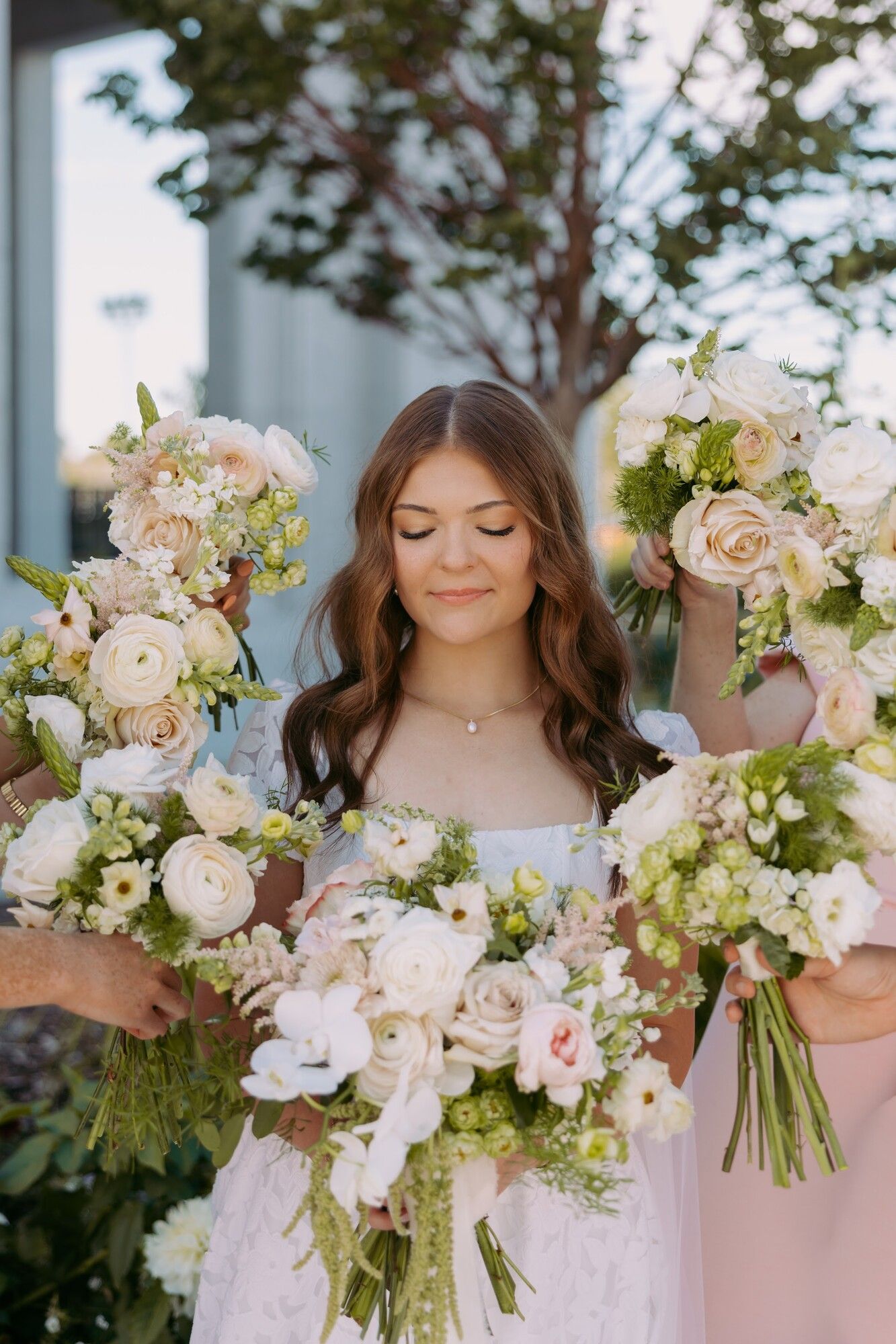 A bride with eyes lowered holding two lush white and blush bouquets under a tree, bridesmaids beside her