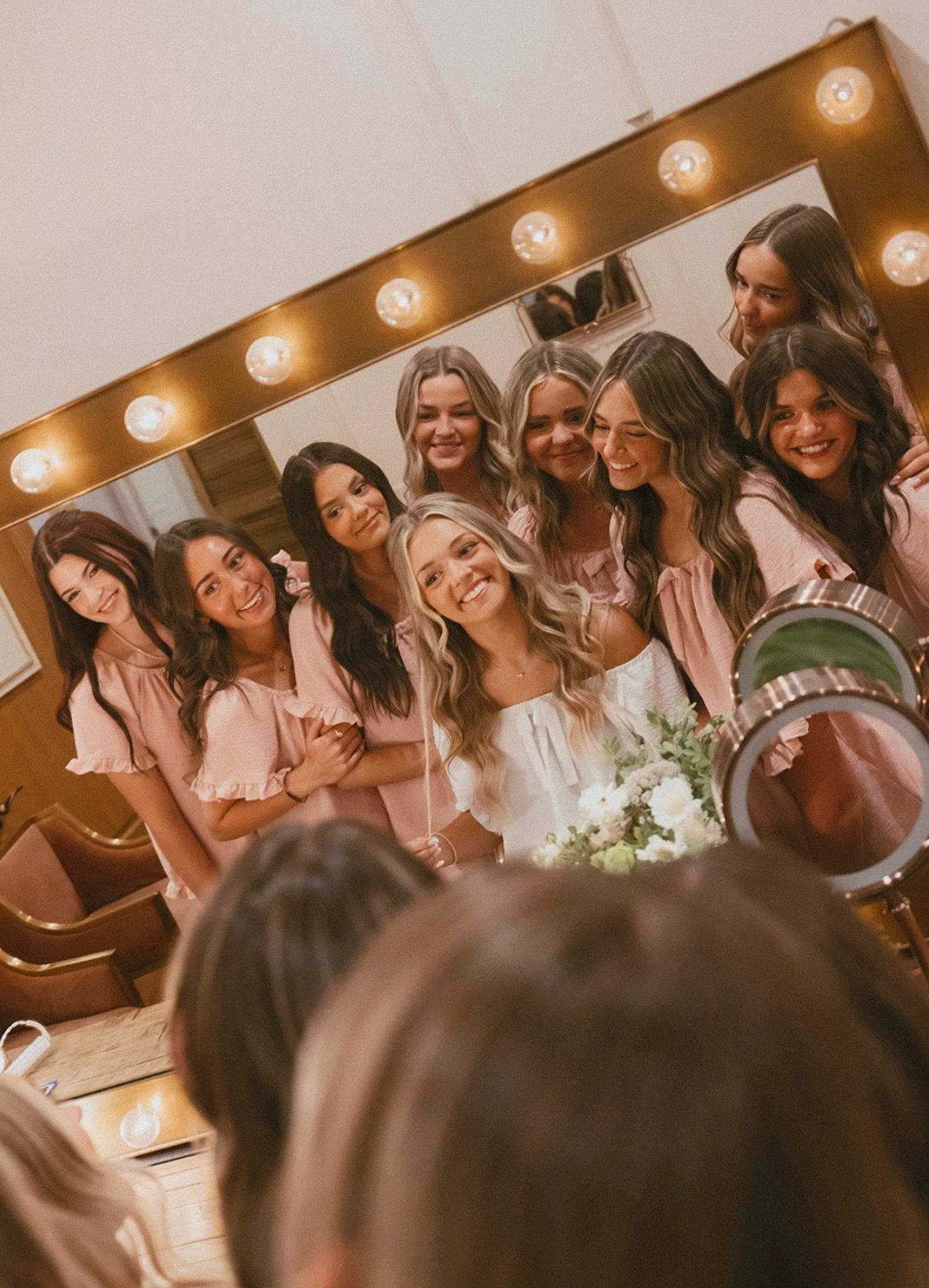 A bride and her bridesmaids crowded around a lit vanity mirror, smiling at their reflection