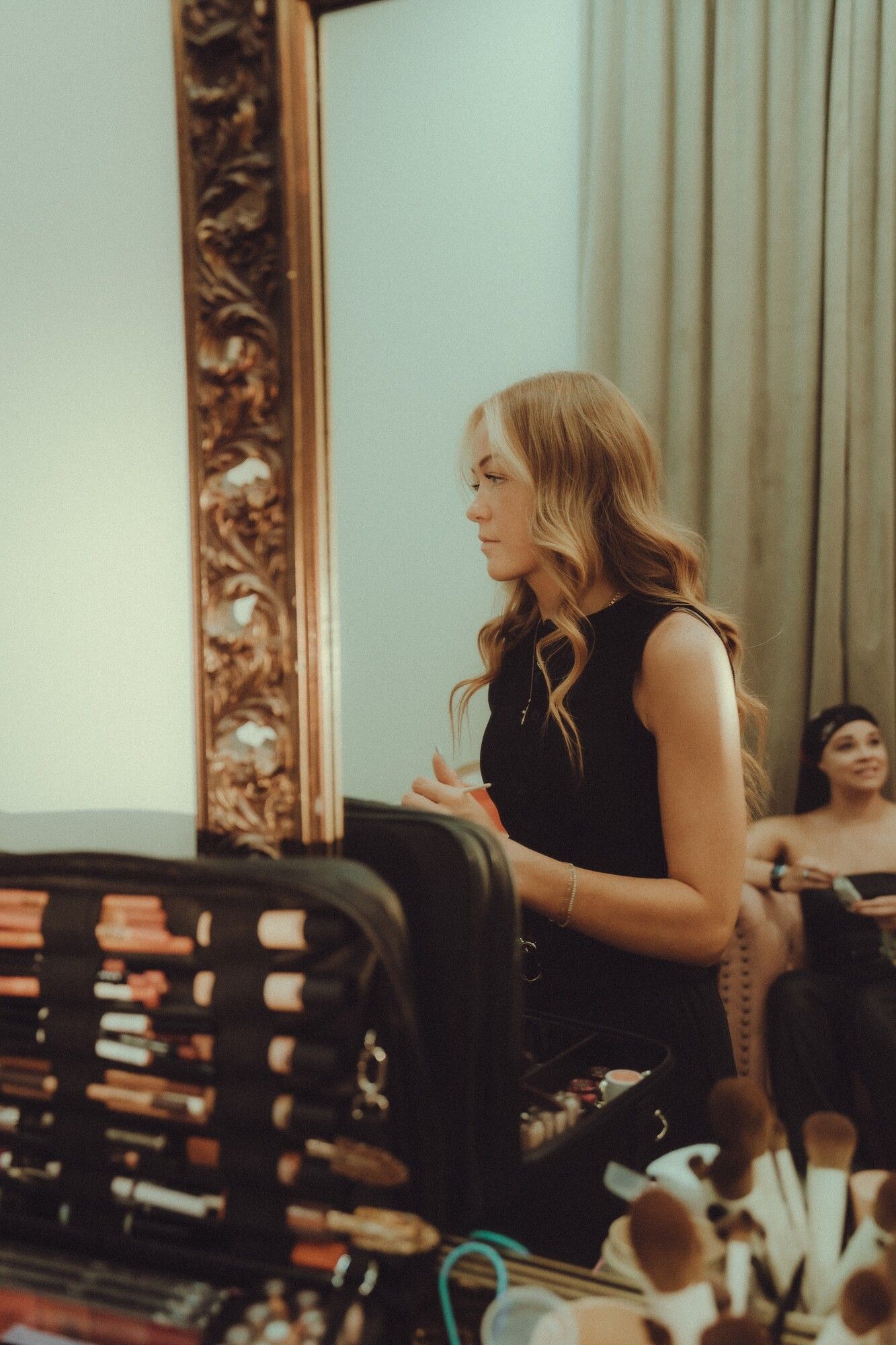 Brynlie at an ornate mirror with her makeup kit open, a client waiting behind her