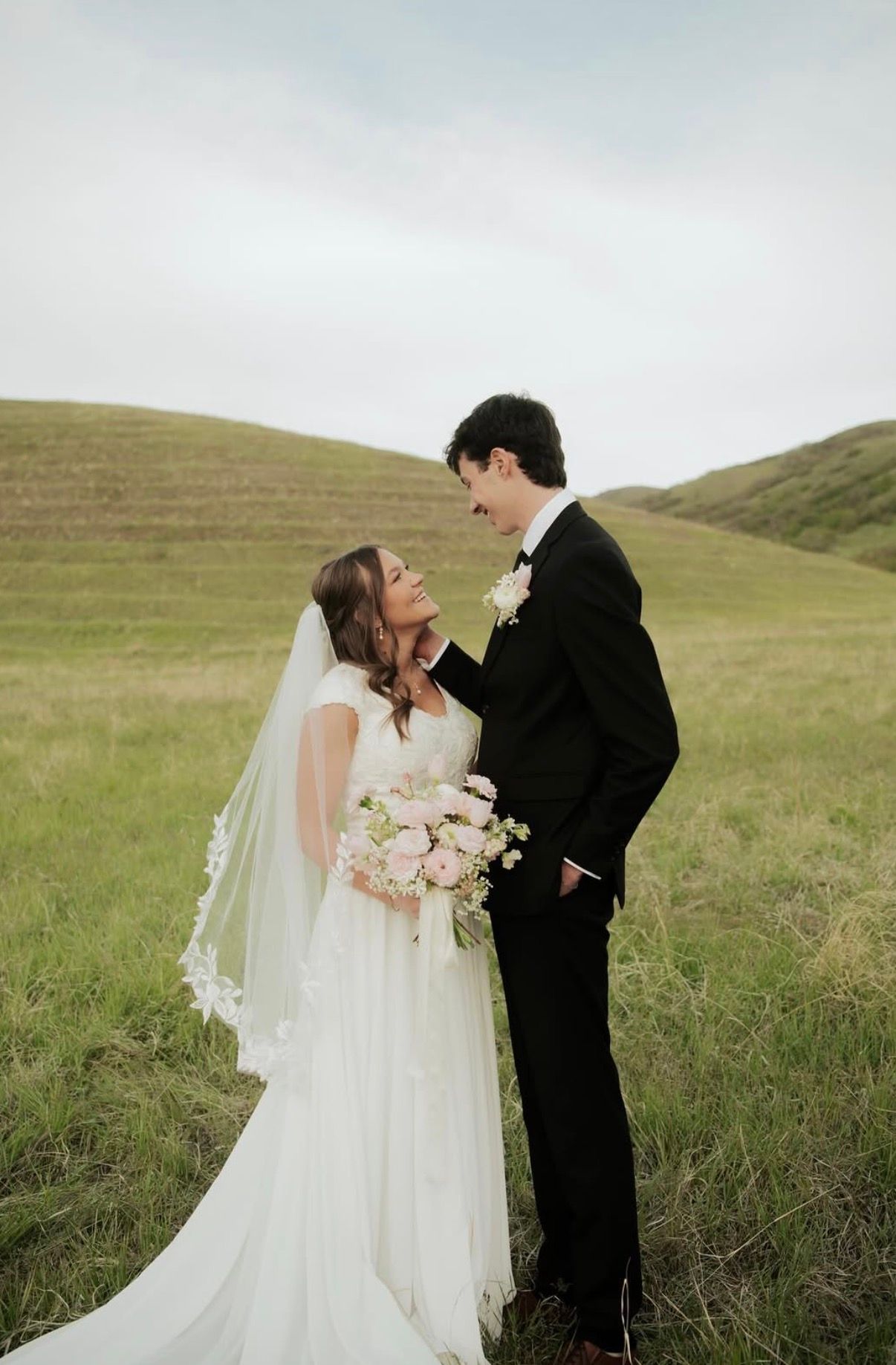 A bride in a veil holding a soft bouquet, looking up at her groom in rolling green hills