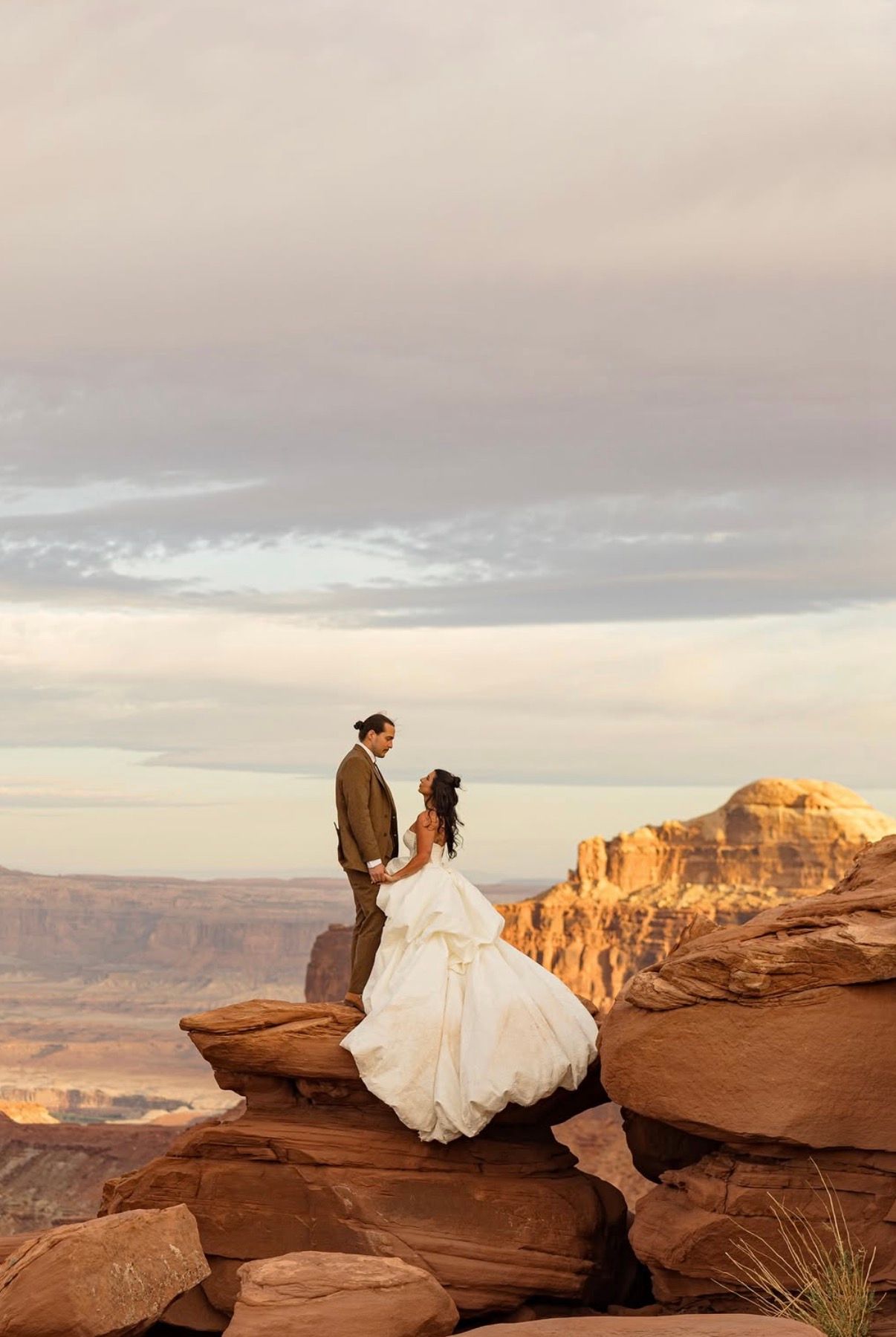 A bride and groom standing on red rock above the Moab desert at dusk, her gown catching the wind