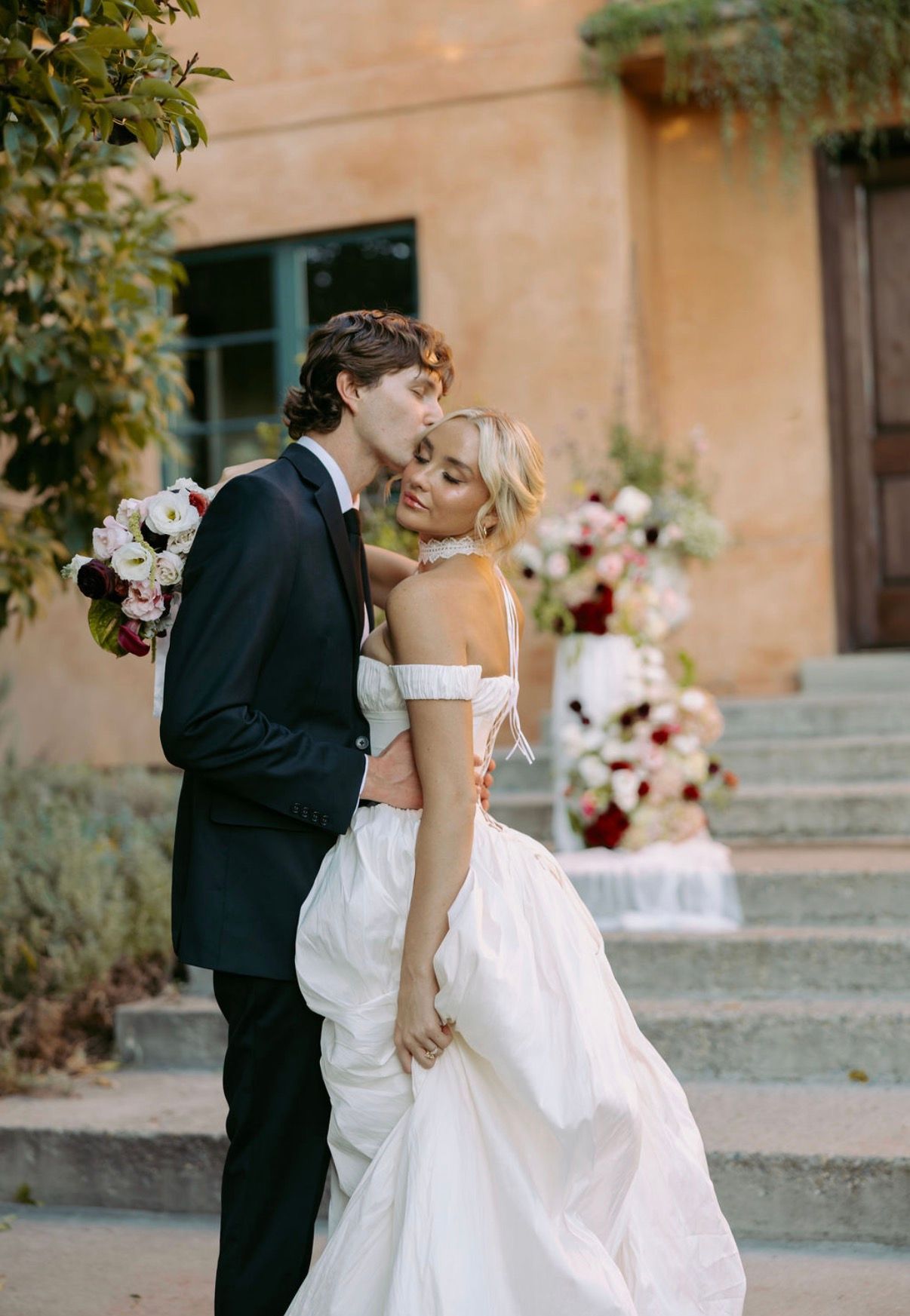 A groom kissing the bride on stone steps lined with white and burgundy florals