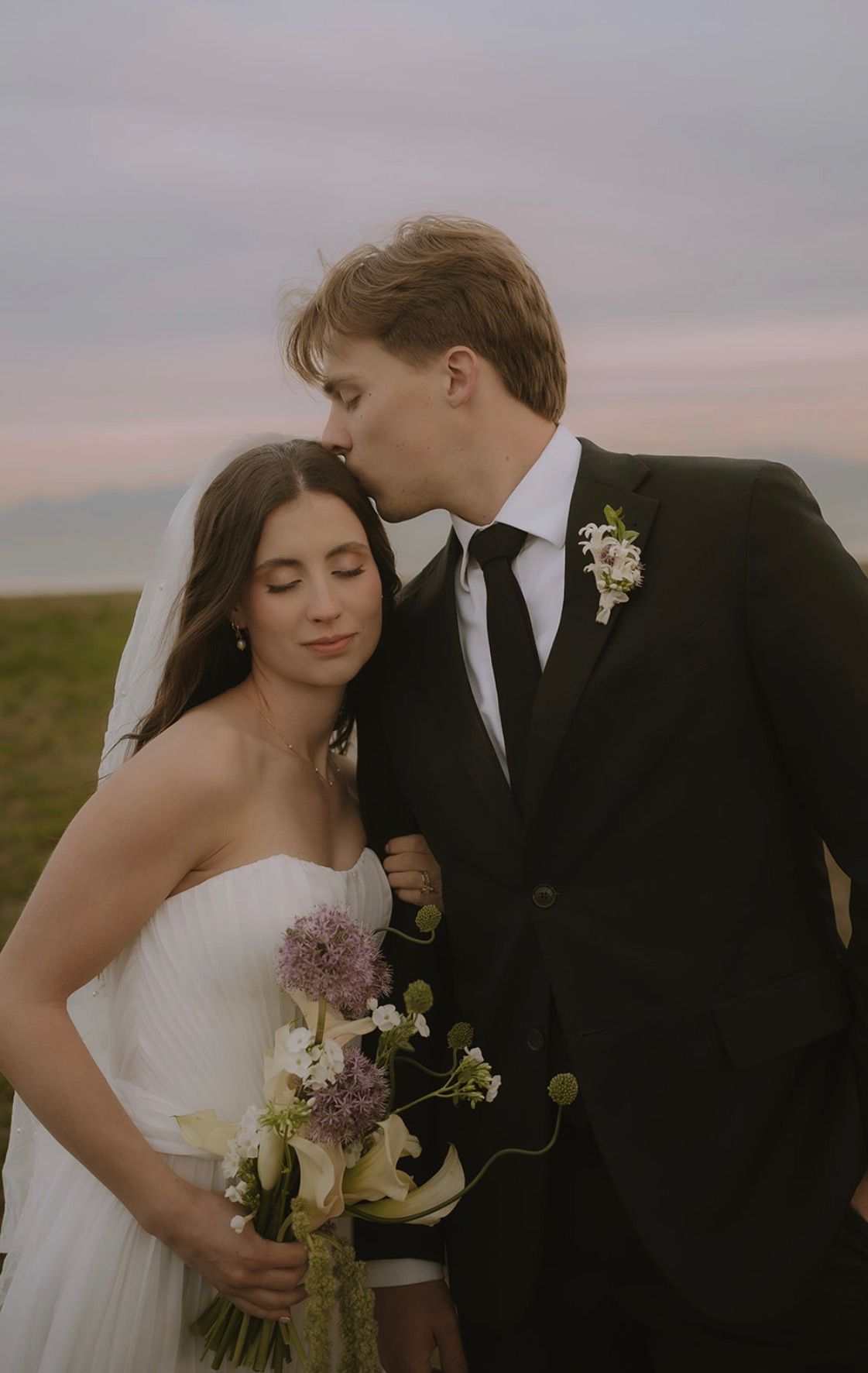 A groom kissing the bride's forehead at sunset, her bouquet of wildflowers in hand