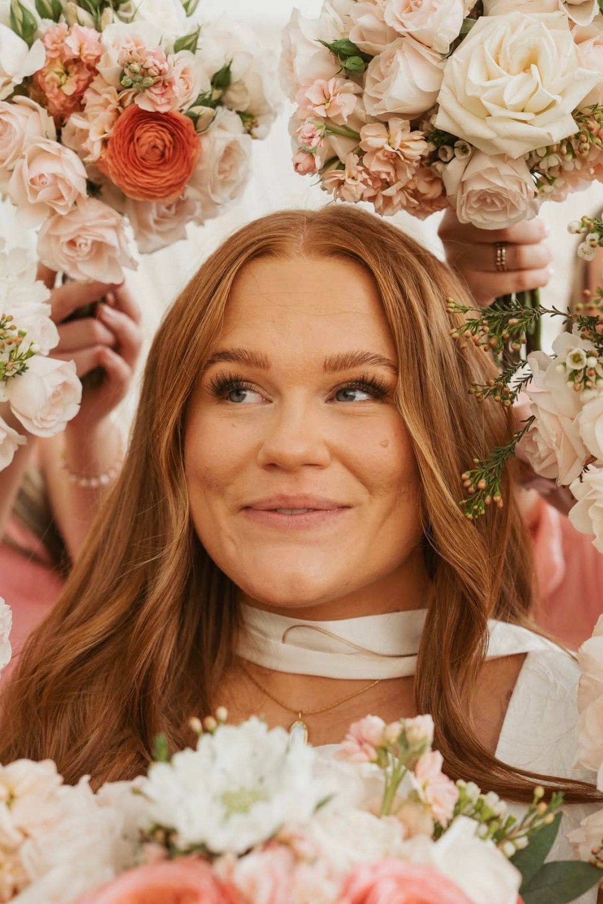 A red-haired bride framed by pale roses, smiling, in soft glam makeup
