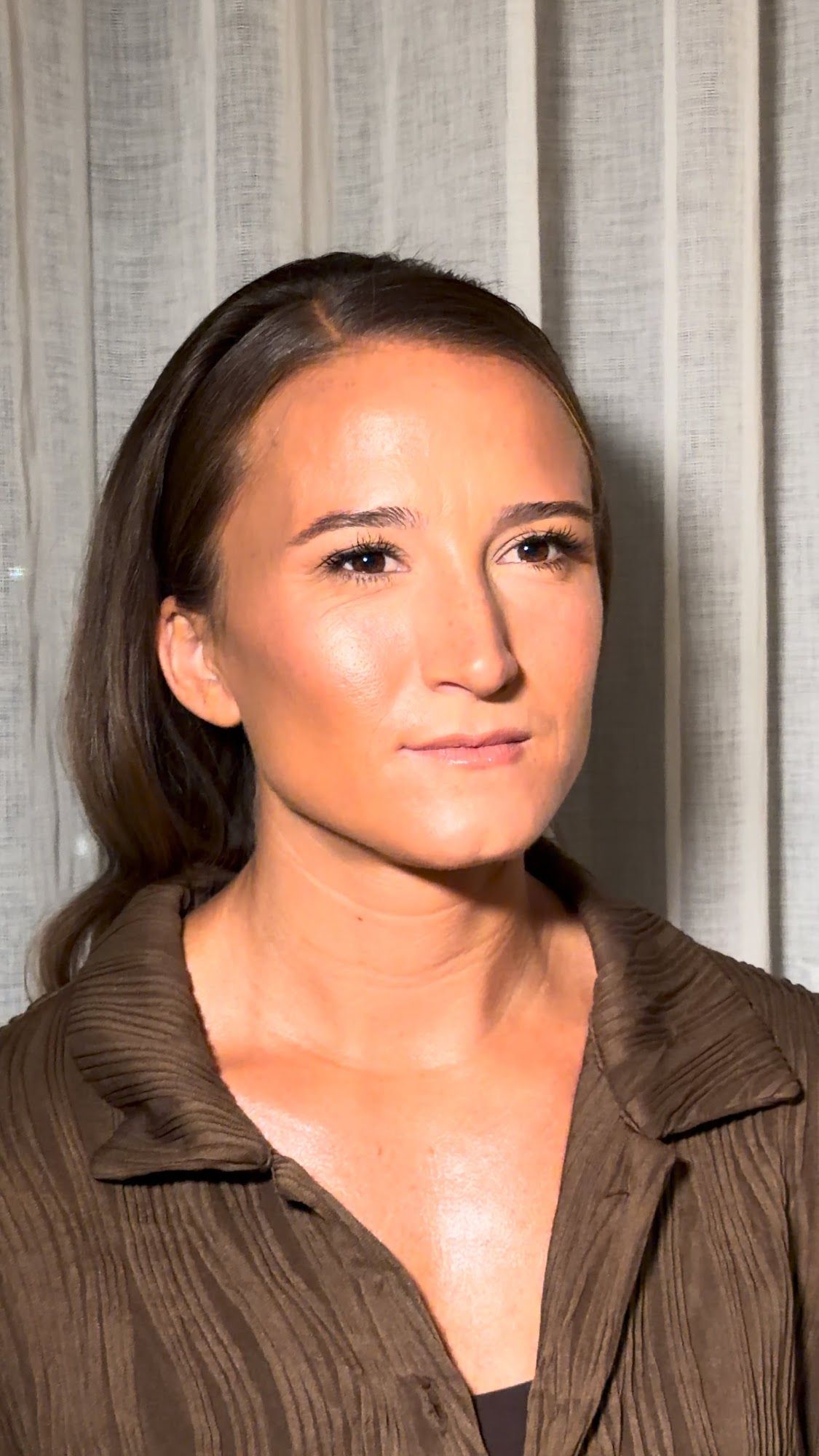 A woman with dark hair pulled back and finished soft glam makeup, in a brown top