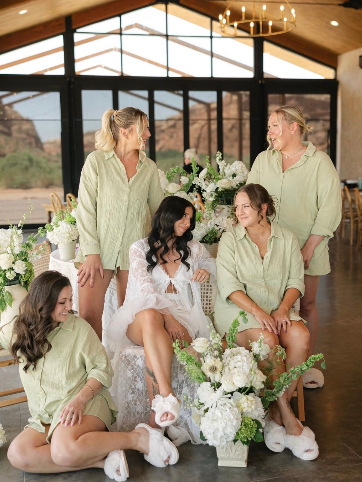 A bride in a lace robe surrounded by her bridesmaids in sage robes, red rock through the windows behind them